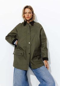 Veste vert olive avec un col en velours côtelé noir, des boutons-pression et deux poches avant ; coupe décontractée avec une texture lisse et des manches raglan.