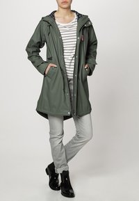 Femme portant un imperméable vert à capuche par-dessus une chemise blanche à rayures, un jean gris clair et des bottines de pluie noires, debout les mains dans les poches.