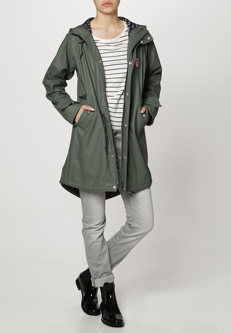 Femme portant un imperméable vert à capuche par-dessus une chemise blanche à rayures, un jean gris clair et des bottines de pluie noires, debout les mains dans les poches.