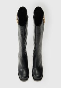 Črne usnjene škornje do kolen s kvadratnimi prsti in z zlatimi dekorativnimi gumbi na zunanjih straneh, gledane z vrha na belem ozadju.