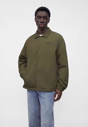 Ung mand iført en olivengrøn jakke med knapper og elastiske manchetter samt blå jeans, der står foran en ensfarvet grå baggrund.