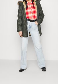 Kobieta w zielonej parze z futrzanym wykończeniem, czerwonej koszuli w kratę, jasnoniebieskich jeansach z wysokim stanem i czarnym paskiem oraz czarnych butach, stojąca z jedną ręką w kieszeni.