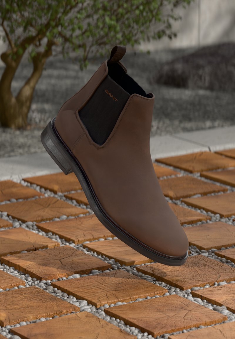 Hnědé kožené chelsea boty s černými elastickými bočními panely, zaoblenou špičkou a nízkým podpatkem, umístěné na texturovaném dřevěném a kamenivovém pozadí.