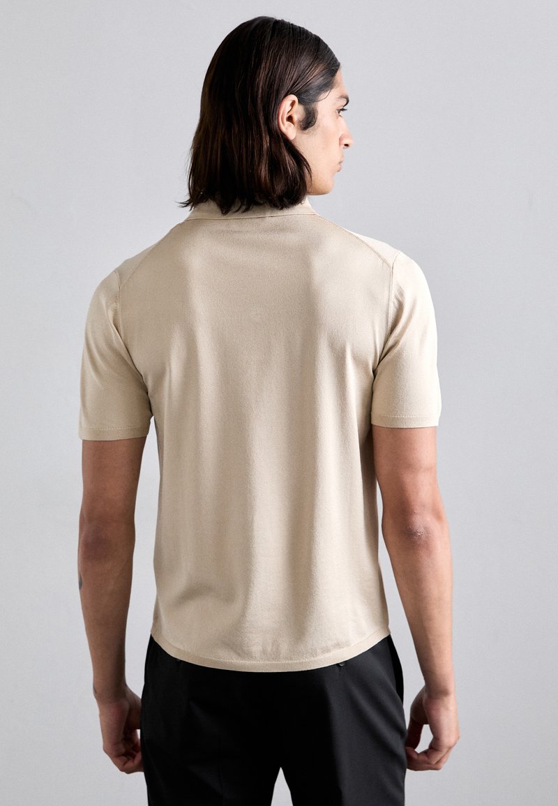 Beige kortärmad polo med mjuk textur. Har en klassisk krage och rundad nederkant. Bärs med mörka byxor, sedd bakifrån.