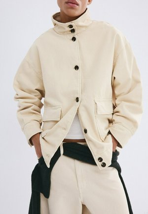 Persona con una chaqueta beige abotonada con grandes bolsillos frontales, manos en los bolsillos y un suéter negro atado alrededor de la cintura.