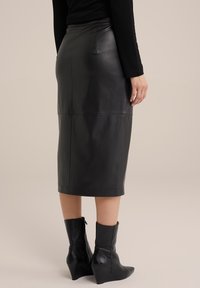 Ženska v črni usnjeni midi svinčnik krilu, črnem dolgih rokavih topu in črnih ankle škornjih z wedges peto, stoji na bež ozadju.