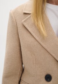 Beige kabát ze směsi vlny s klopami, dvouřadým zapínáním a černými knoflíky. Má jemnou strukturu a boční kapsy.