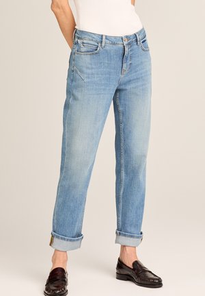 Hellblaue, aufgerollte Jeans, getragen mit schwarzen Loafers, von der Taille bis zu den Knöcheln vor neutralem Hintergrund gezeigt.