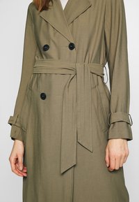 Oliwkowy płaszcz trench z podwójnymi czarnymi guzikami, paskiem w talii, bocznymi kieszeniami i regulowanymi paskami na mankietach obu rękawów.