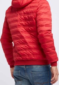 Röd vadderad jacka med horisontell kviltad mönster, justerbar huva och elastiska ärmslut. Matchad med denimjeans.