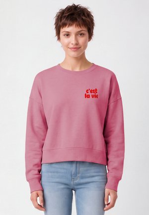 Junge Person mit kurzen braunen Haaren, die einen pinken Sweatshirt mit rotem "c'est la vie" Text und hellblauen Jeans trägt, vor einem einfarbigen Hintergrund steht.
