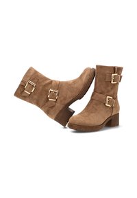 Las botas de tobillo de ante marrón cuentan con dos hebillas ajustables, un tacón apilado y una punta redondeada. La textura es suave, lo que realza su diseño moderno.