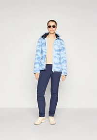 Light blue cloud-patterned jacket over a beige top, paired with dark navy pants and light-colored footwear. Features a hood and sunglasses.