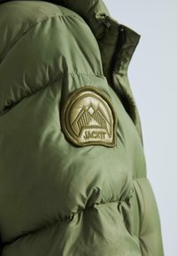 Olīvu zaļa pūkaina jaka ar spīdīgu tekstūru. Izceļas ar izteiktu apaļu uzšuvi ar kalnu grafiku un tekstu "JACKIT" zeltā.