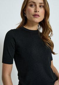 Top de punto negro de manga corta con cuello redondo, que presenta bordes acanalados y una textura suave. Sin patrones ni adornos visibles.