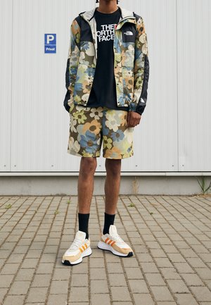 Henkilö, joka pukeutuu kukkakuviolliseen takkiin ja shortseihin, mustaan The North Face -t-paitaan, mustiin sukkiin ja valkoisiin Adidas-tennareihin, joissa on oranssit raidat.