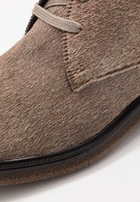 Botte de cheville en fourrure brune avec un dessus texturé, une fermeture à lacets et des coutures noires le long de la semelle, avec une finition douce et semblable à du daim.