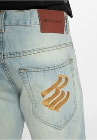 Heledad sinise denimi teksad, millel on tekstureeritud pind, sisaldades pruuni naharatset, millel on silt "ROCAWEAR" ja kuldne tikand tagakotil.