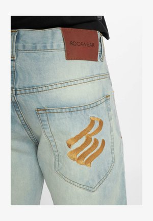 Lyseblå denim jeans med en struktureret overflade, der har et brunt lædermærke mærket "ROCAWEAR" og et guld broderet design på baglommen.