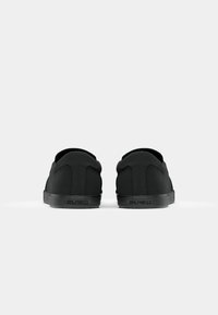 Paire de baskets noires sans lacets vues de dos, avec semelles en caoutchouc texturées et nom de marque embossé sur la languette du talon.