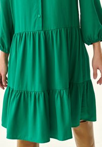 Vestido verde de volantes en capas, hecho de una tela suave, con cierre de botones y mangas de globo, que presenta volantes fruncidos en el dobladillo y en los puños.