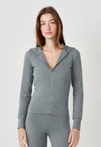 Joven mujer con cabello largo y rojo, vistiendo una sudadera con capucha gris con cremallera y leggins a juego, de pie contra un fondo claro y sencillo.