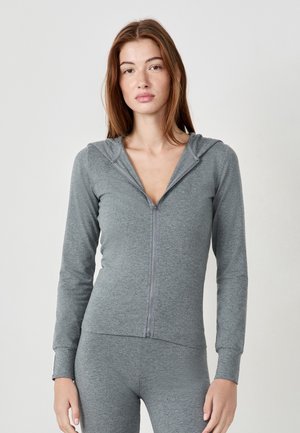 Junge Frau mit langen roten Haaren, die einen grauen Kapuzenpullover mit Reißverschluss und passende Leggings trägt, steht vor einem schlichten hellen Hintergrund.
