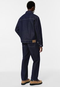 Giacca in denim blu scuro con pantaloni abbinati, presenta una toppa in pelle marrone e cuciture a contrasto. Calzature beige con suole nere.