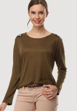 Joven mujer con cabello castaño en una coleta baja que lleva una camiseta de manga larga verde oliva y pantalones beige claros, sonriendo con la mano en la cadera.
