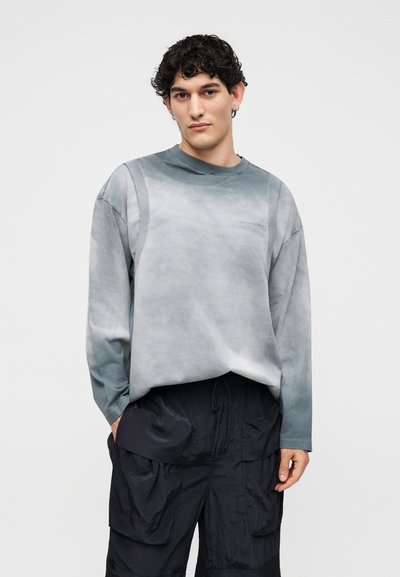 Νέος άνδρας με σγουρά μαλλιά, που φοράει ξεθωριασμένο γκρι πουκάμισο με μακριά μανίκια και μαύρο παντελόνι cargo, στέκεται μπροστά σε ένα απλό φωτεινό φόντο.