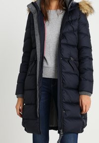 Marinblå puffjacka med en foder av fuskpäls, quiltat mönster, dragkedjestängning, sidofickor och grå stickade manschetter. Bärs över en grå tröja.