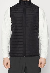 Schwarze gepolsterte Weste mit vertikalen Steppmuster, hohem Kragen, vorne mit Reißverschluss und zwei Seitentaschen, getragen über einem weißen Langarmshirt.