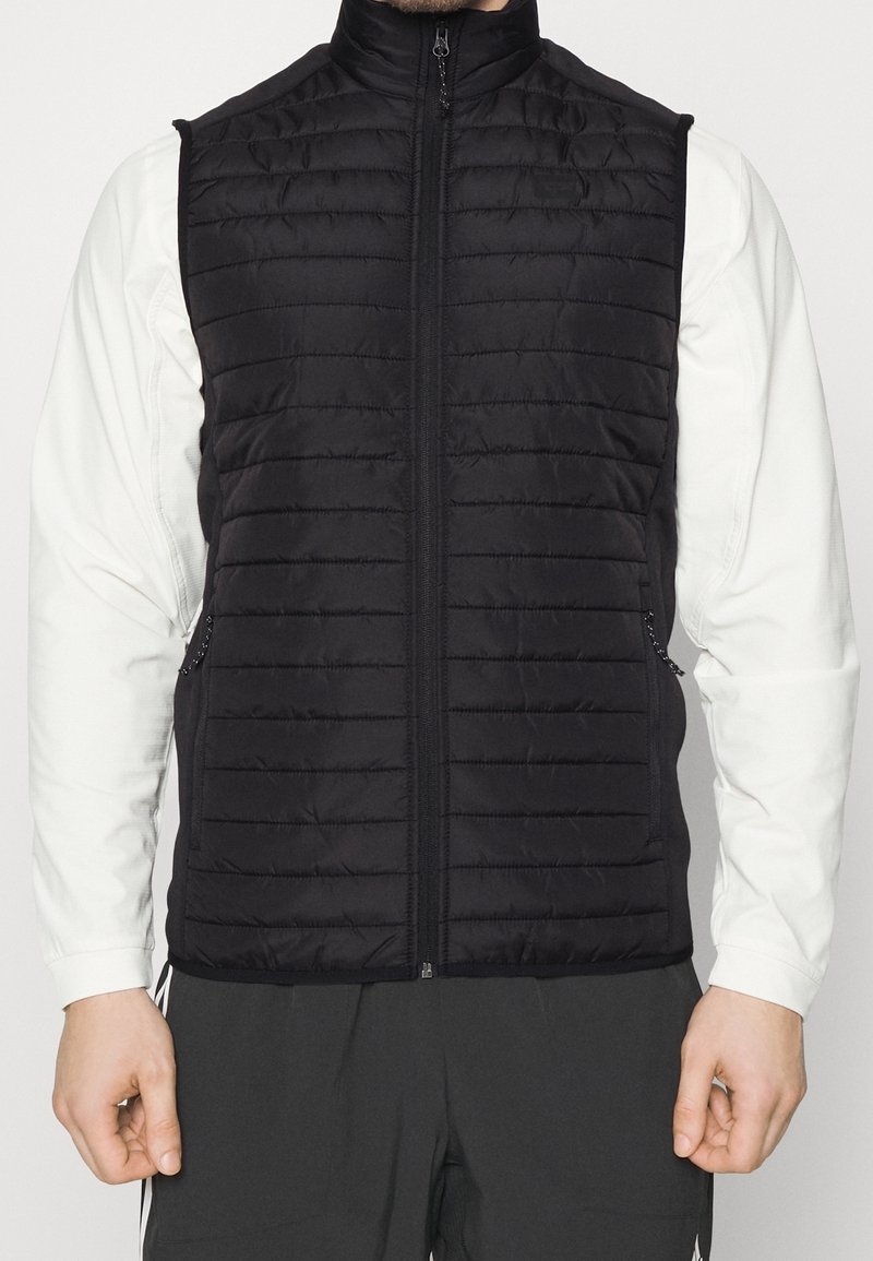 Schwarze gepolsterte Weste mit vertikalen Steppmuster, hohem Kragen, vorne mit Reißverschluss und zwei Seitentaschen, getragen über einem weißen Langarmshirt.