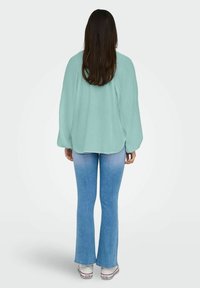 En ljus teal oversized skjorta med långa ärmar, texturerad tyg och rundad nederkant, ihop med utsvängda blå jeans och vita canvas sneakers.