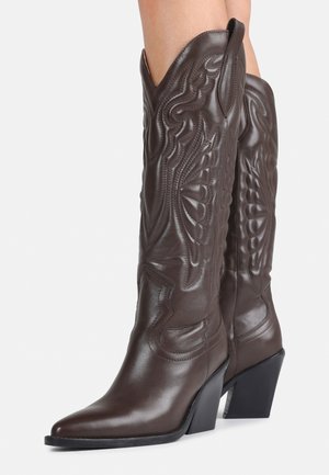 Mørkebrune cowboyboots i skinn opp til kneet med spiss tupp, dekorative sømmer og skrå blokkhæl, båret av en person.