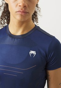 Camisola atlética azul-marinho com padrões de malha sutis, detalhes em laranja e um logótipo no lado esquerdo do peito, feita de um tecido leve e elástico.