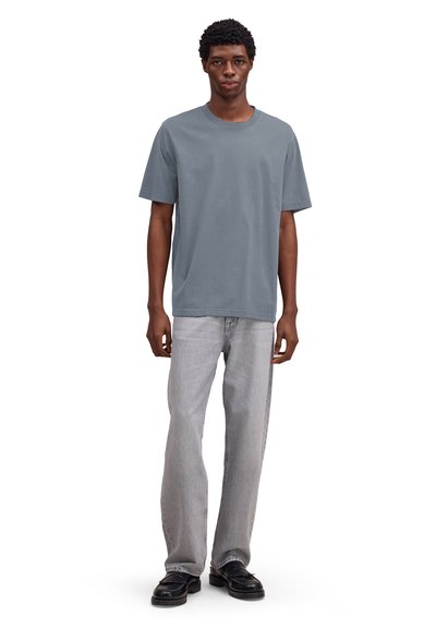 Jeune homme debout, portant un t-shirt gris uni à manches courtes, un jean gris clair et des mocassins noirs, sur un fond blanc.