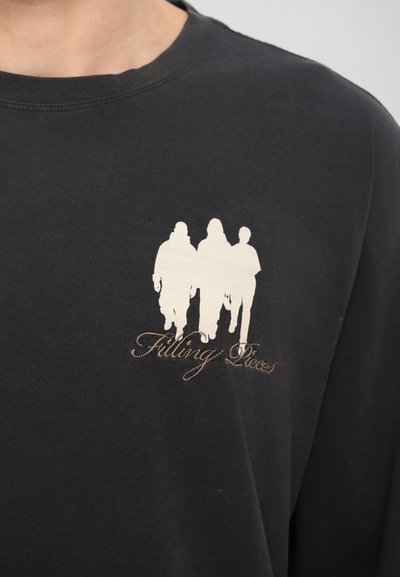 Melnā garām piedurknēm T-krekls ar baltu silueta grafiku, uz kuras attēloti trīs figūras, un apakšā ir teksts "Filling Pieces" izliekta rakstā.