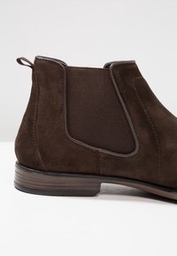 Bottine en suède marron avec panneaux latéraux élastiques et semelle en cuir lisse. Présente des détails de couture et une languette pour faciliter l'enfilage.