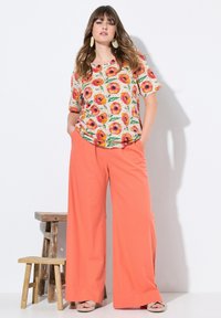 Bloemmotief top met korte mouwen, roomkleurige achtergrond; brede koraalkleurige broek; plateauzolen sandalen; eenvoudige houten kruk op de achtergrond.