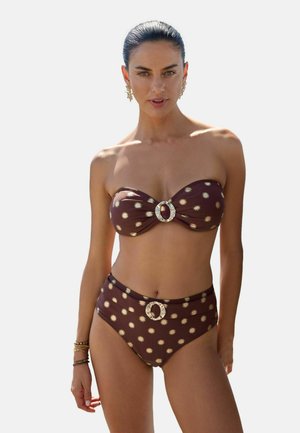 Juuksed tagant siledaks kammitud naine kannab pruuni bandeau-bikinid, millel on päikesemustrid ja dekoratiivsed ümardatud pandlad peal, ning kõrge vöökohaga alumisi pükse.