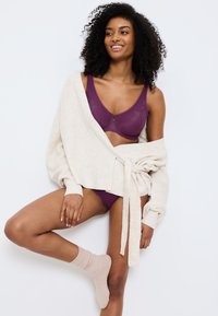 Fialová podprsenka a spodní prádlo sladěné s volným, krémovým žebrovaným svetrem s páskem v pase a strukturovanými rukávy.