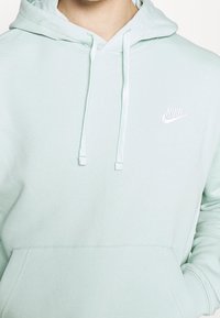 Mintgrön hoodie med dragsnören, känguruficka och vit Nike-logotyp på vänster bröst. Tillverkad av mjukt, texturerat tyg.