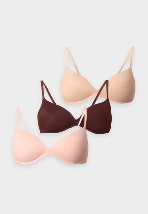 Kvinde står barfodet iført beige bh og højtaljede trusser, ser en smule op og til siden mod en ensfarvet baggrund.