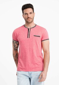 Rød T-shirt med kortærmer, rund halsudskæring, sort-hvid stribet detalje, knaplukning og en lille brystlomme.