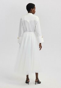 Witte jurk met een aansluitend bovenlijf en een vloeiende tule rok. Lange mouwen, gouden knopelementen op de manchetten en een taille accentuering.