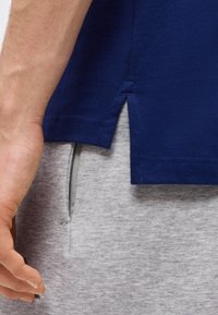 Gros plan sur la main d'une personne près de la fente latérale d'une chemise bleu marine portée avec un pantalon gris clair doté d'une poche sur la couture latérale.