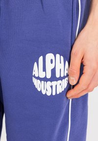 Vijolične trenirke iz bombaža, ki imajo velik bel logotip "ALPHA INDUSTRIES" na stegnu in kontrastno belo obrobo ob straneh.
