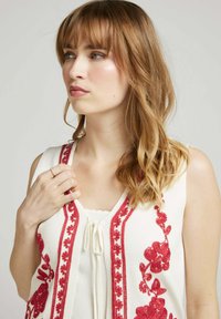 Haut sans manches blanc orné de broderies florales rouges le long des bords avant et d'un cordon de serrage au niveau du col, avec des détails complexes.