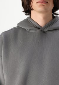 Hanorac gri cu glugă realizat dintr-un material moale, având o textură ribbită și un decolteu adânc. Designul include un stil clasic al glugii.